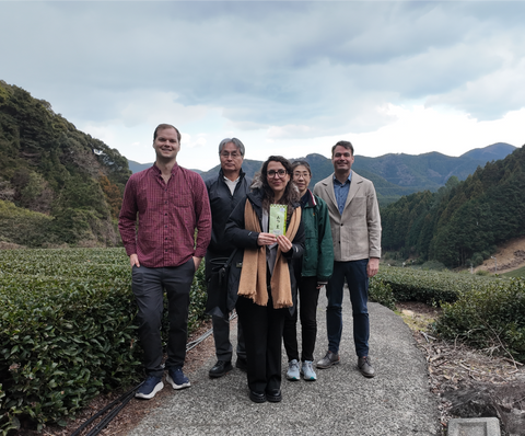 Nio Teas' team and farmers standing together on a gravel path in a mountainous tea plantation setting under an overcast sky.