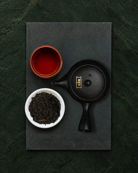 A top-down view of a black metal teapot with a gold emblem, a small white bowl of fermented japanese tea, and a red cup, all arranged on a dark slate tray over a dark green textured background.