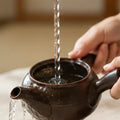 A close-up of a dark brown speckled ceramic teapot with water being poured into it from a twisted metal infuser strainer, held by hands in a softly blurred indoor setting.